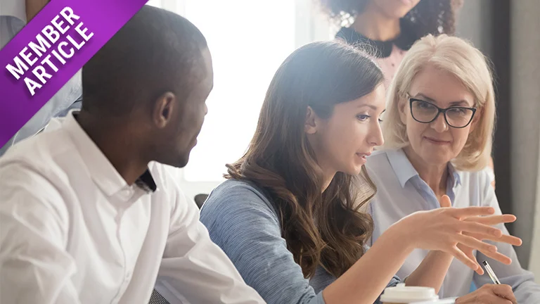 Stock image of a dark-haired white woman at profile view, in mid-discussion with her colleagues sitting down at a table: a black man in a white collared shirt to her right, and a middle-aged white woman with blonde hair and glasses to her left (both of whom are looking at her).
